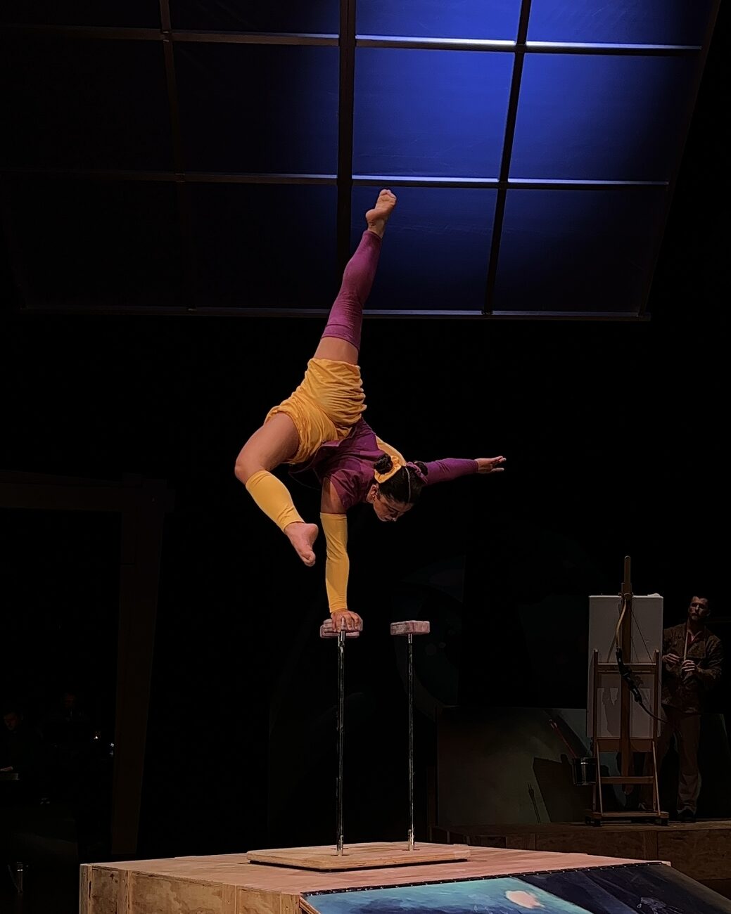 Anne Arellano in a one-arm handstand on canes in a yellow and purple costume, theatre stage.