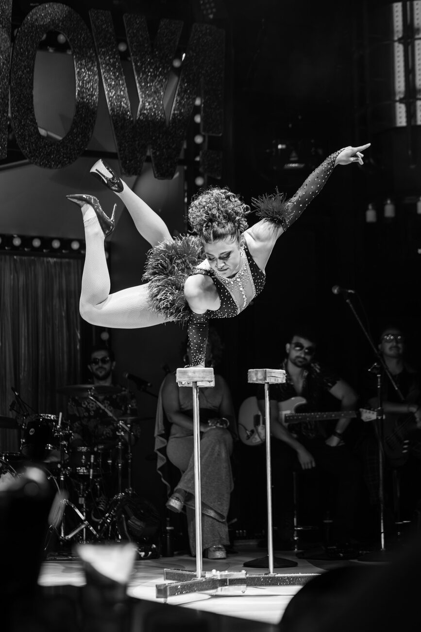 Black and white photo of Anne Arellano in a one-arm handstand on canes with a live band behind her.