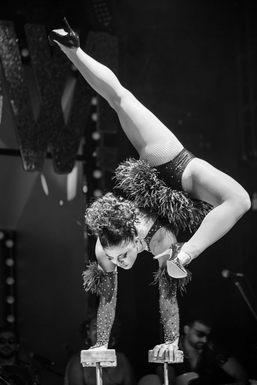 Black and white photo of Anne Arellano in a split handstand on canes, feathered costume and heels.