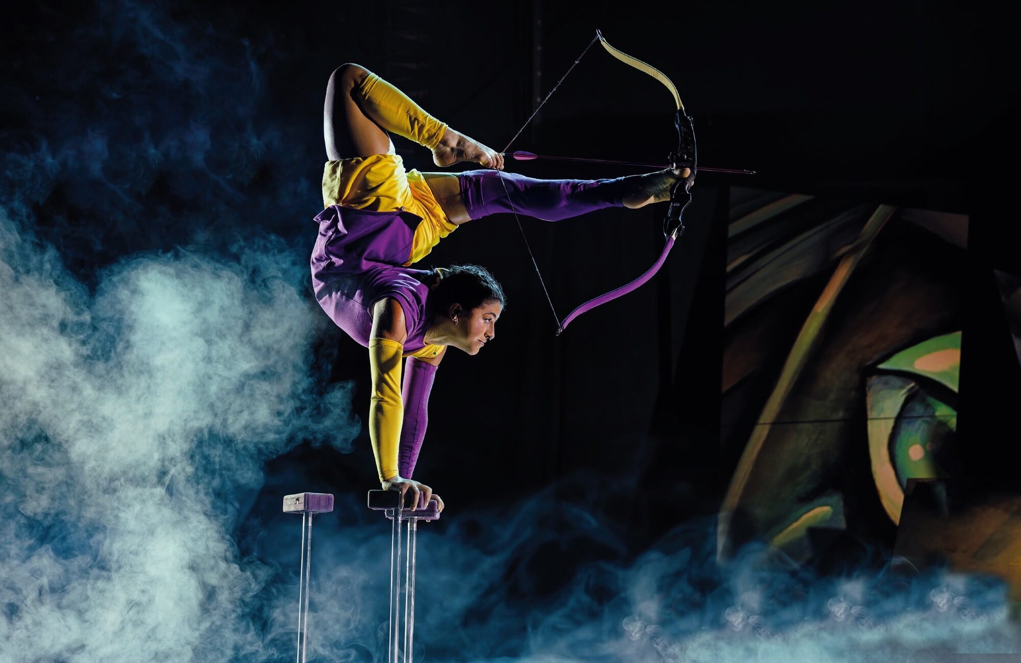 Anne Arellano drawing a bow with her feet in a one-arm handstand, stage smoke around her.